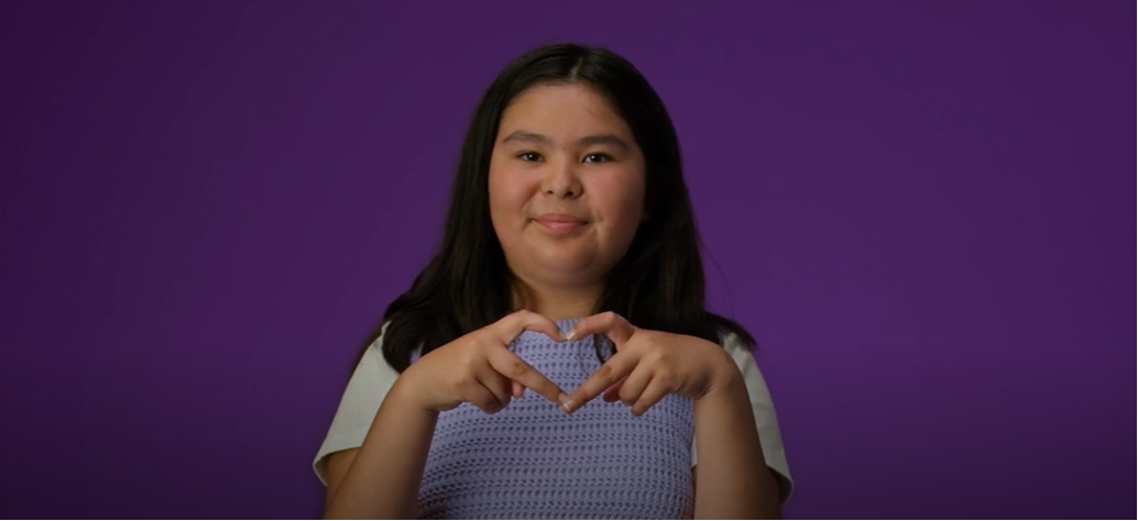 A young person making a heart with their hands against a purple backdrop
